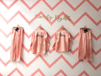 A couple of pink satin pajama sets hanging on hangers against a chevron patterned wall, with the names 'Danny' and 'Claire' printed on the back of the shirts.